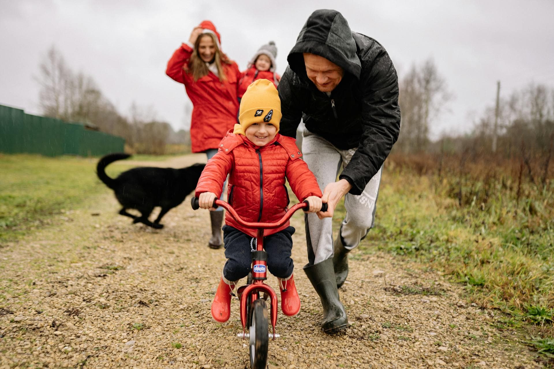 Dad pushes son outside with hes son on the red bike and mom holds the baby the mom and kids waring red jackets.