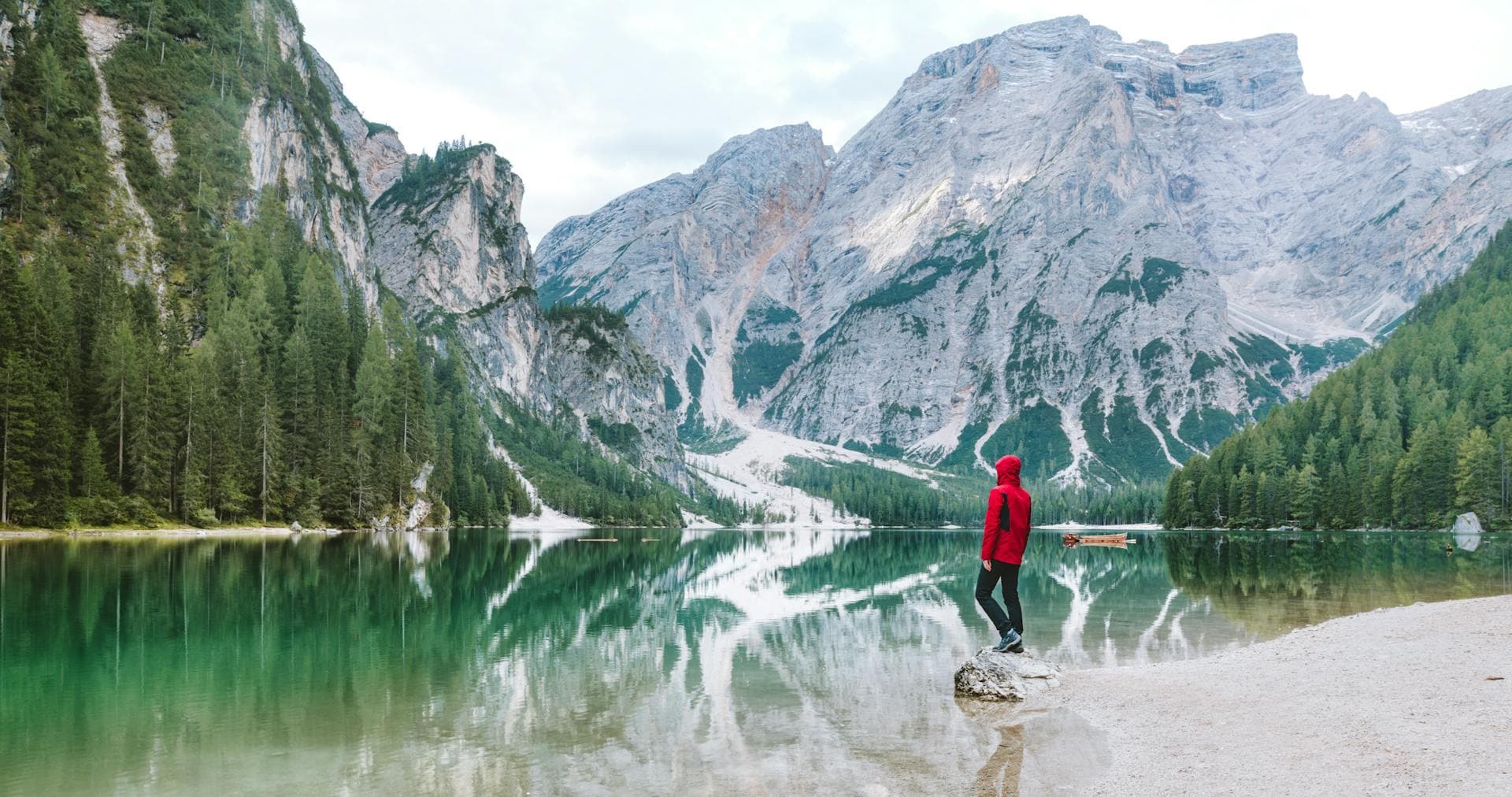 Man standing on a stony beach surrounded with green pine trees and snowy mountains