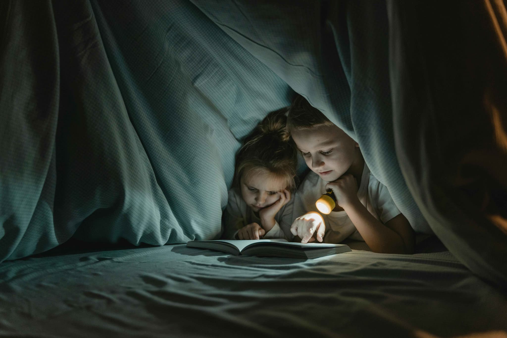 Kids reading a book with a flashlight inside a self made tent of blankets